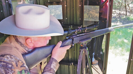 Man aiming a scoped rifle while wearing a cowboy hat.