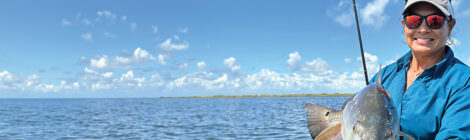 A fish being held over the water under a blue sky with clouds.