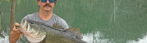 Man holding a large fish by a lake, wearing sunglasses and a cap.