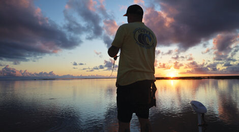 A person fishing at sunset by a calm lake with mountains in the background.