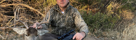 Hunter posing with a deer and rifle in a wooded area.