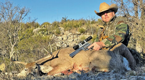 Hunter poses with a deer in a dry, rugged landscape under clear blue skies.