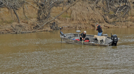 Two people fishing from a small boat on a calm river near dry, leafless trees.